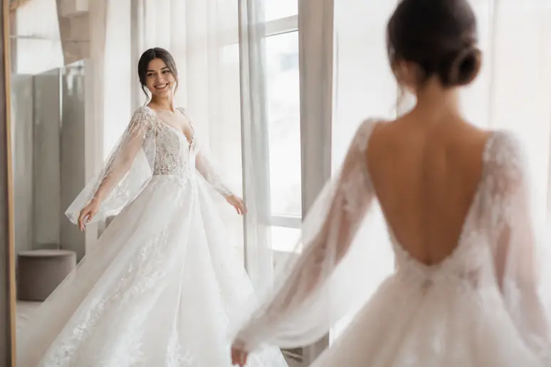 Une mariée rayonnante essaie une robe de princesse volumineuse aux détails de dentelle, admirant son reflet dans un espace d'essayage élégant.