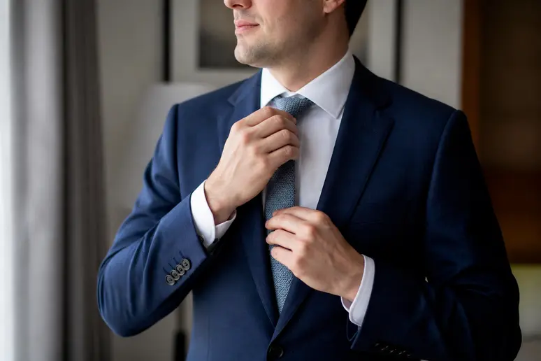 Un homme ajuste sa cravate en soie sur une chemise blanche avec son costume mariage homme bleu.