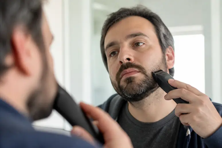 Homme taillant sa barbe de trois jours avec une tondeuse électrique devant le miroir